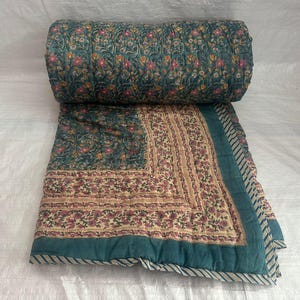 Peut inclure: Une couverture matelassée présentant un fond bleu sarcelle foncé avec un motif floral dense en rose, orange et blanc. La partie inférieure affiche une bordure contrastante crème avec un imprimé floral plus petit et un bord rayé bleu et blanc.