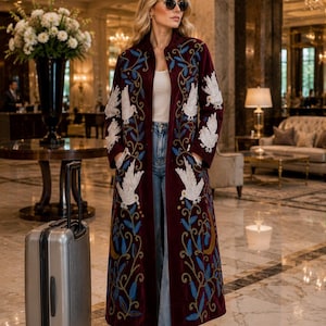 May include: A long, deep burgundy velvet coat featuring intricate white bird embroidery and blue leaf patterns. The coat is worn over a white top and light blue denim jeans, paired with a silver rolling suitcase standing on the floor nearby.