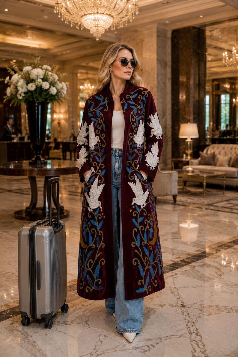 May include: A long, deep burgundy velvet coat featuring intricate white bird embroidery and blue leaf patterns. The coat is worn over a white top and light blue denim jeans, paired with a silver rolling suitcase standing on the floor nearby.