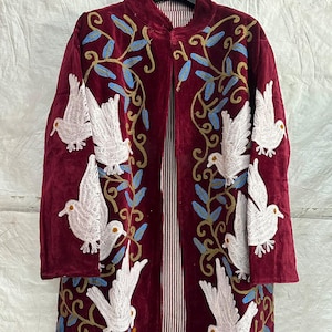 May include: A long-sleeved, open-front coat in deep burgundy velvet, featuring intricate embroidery of white doves and blue leafy vines. The interior lining is visible, showcasing a vertical red and white striped pattern throughout the garment.