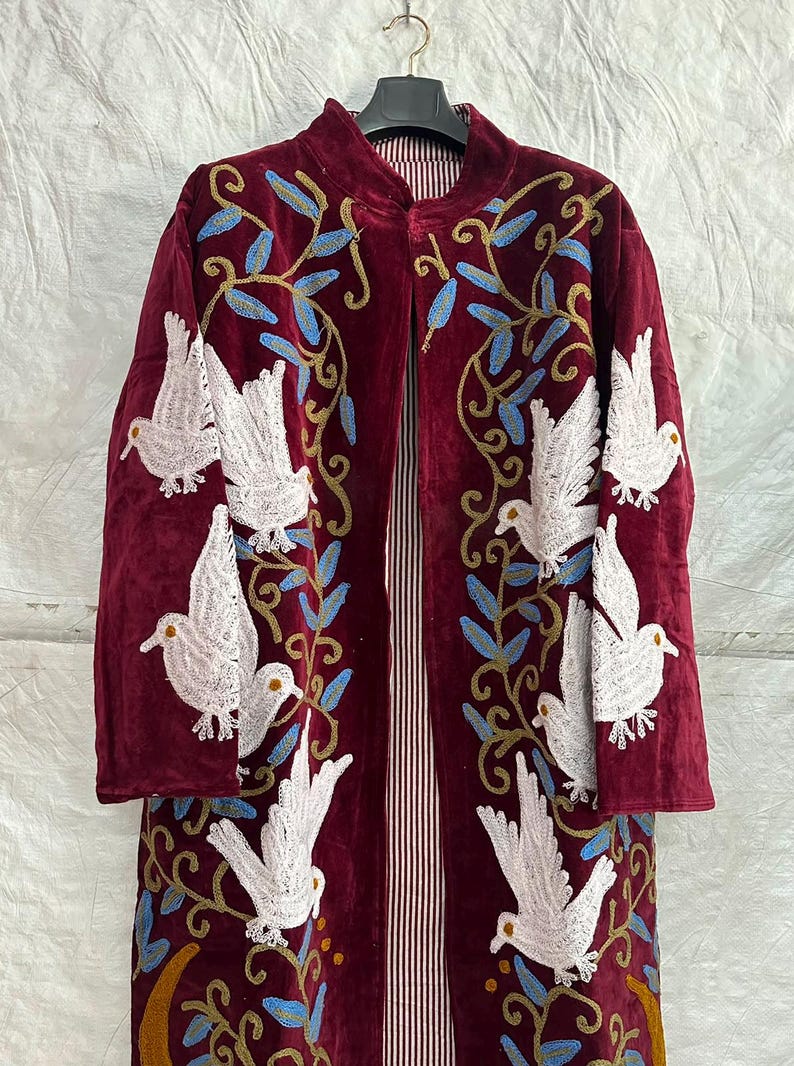May include: A long-sleeved, open-front coat in deep burgundy velvet, featuring intricate embroidery of white doves and blue leafy vines. The interior lining is visible, showcasing a vertical red and white striped pattern throughout the garment.