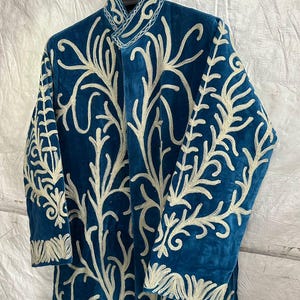May include: A long-sleeved, teal blue velvet coat featuring an intricate, cream-colored embroidered pattern that covers the front and sleeves. The garment has a high, structured collar and is displayed on a black hanger against a white background.