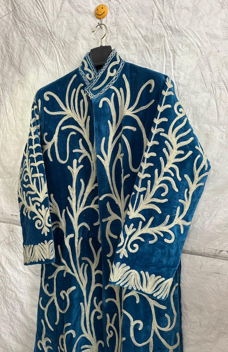 May include: A long-sleeved, teal blue velvet coat featuring an intricate, cream-colored embroidered pattern that covers the front and sleeves. The garment has a high, structured collar and is displayed on a black hanger against a white background.