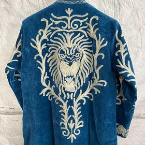 May include: A long-sleeved blue velvet coat featuring intricate white embroidery on the back. The design displays a large, detailed lion head surrounded by ornate, swirling floral patterns that extend from the collar down to the lower back area.