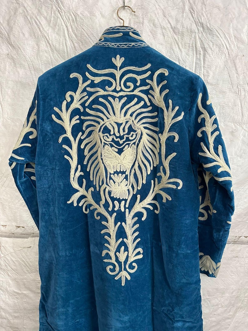 May include: A long-sleeved blue velvet coat featuring intricate white embroidery on the back. The design displays a large, detailed lion head surrounded by ornate, swirling floral patterns that extend from the collar down to the lower back area.