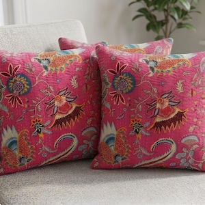 May include: Two square throw pillows featuring a vibrant hot pink base color with a multi-colored floral and paisley pattern. The fabric displays intricate stitching details throughout, showcasing various stylized botanical motifs in shades of orange, blue, yellow, and dark red.