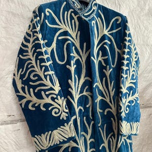 May include: A long-sleeved, deep blue velvet coat featuring an intricate, light-colored embroidered pattern that covers the front and sleeves. The garment has a high, structured collar and is displayed on a black hanger against a white background.