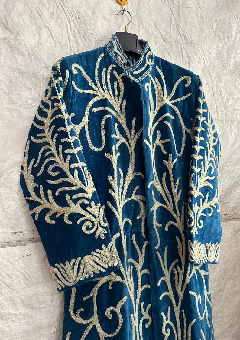 May include: A long-sleeved, deep blue velvet coat featuring an intricate, light-colored embroidered pattern that covers the front and sleeves. The garment has a high, structured collar and is displayed on a black hanger against a white background.