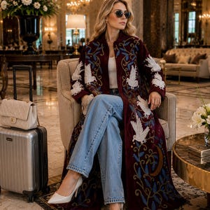 May include: A long, deep burgundy coat featuring intricate white bird embroidery and blue leaf patterns, worn over a white top and light blue denim jeans. The ensemble is paired with pointed-toe cream heels, a cream quilted handbag, and a silver suitcase.