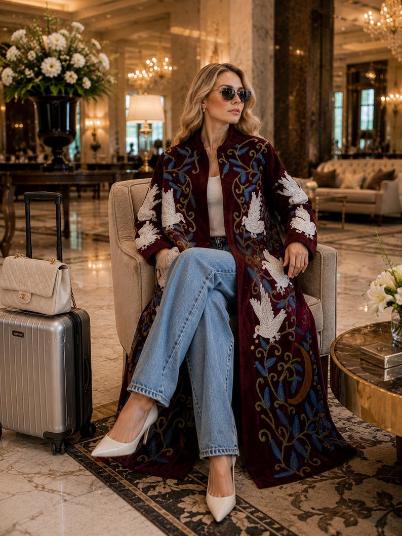 May include: A long, deep burgundy coat featuring intricate white bird embroidery and blue leaf patterns, worn over a white top and light blue denim jeans. The ensemble is paired with pointed-toe cream heels, a cream quilted handbag, and a silver suitcase.