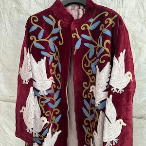 May include: A deep burgundy velvet coat featuring intricate embroidery of white doves and blue leafy vines. The garment has a high collar, long sleeves, and a vertical striped lining visible at the front opening, all displayed on a black hanger.