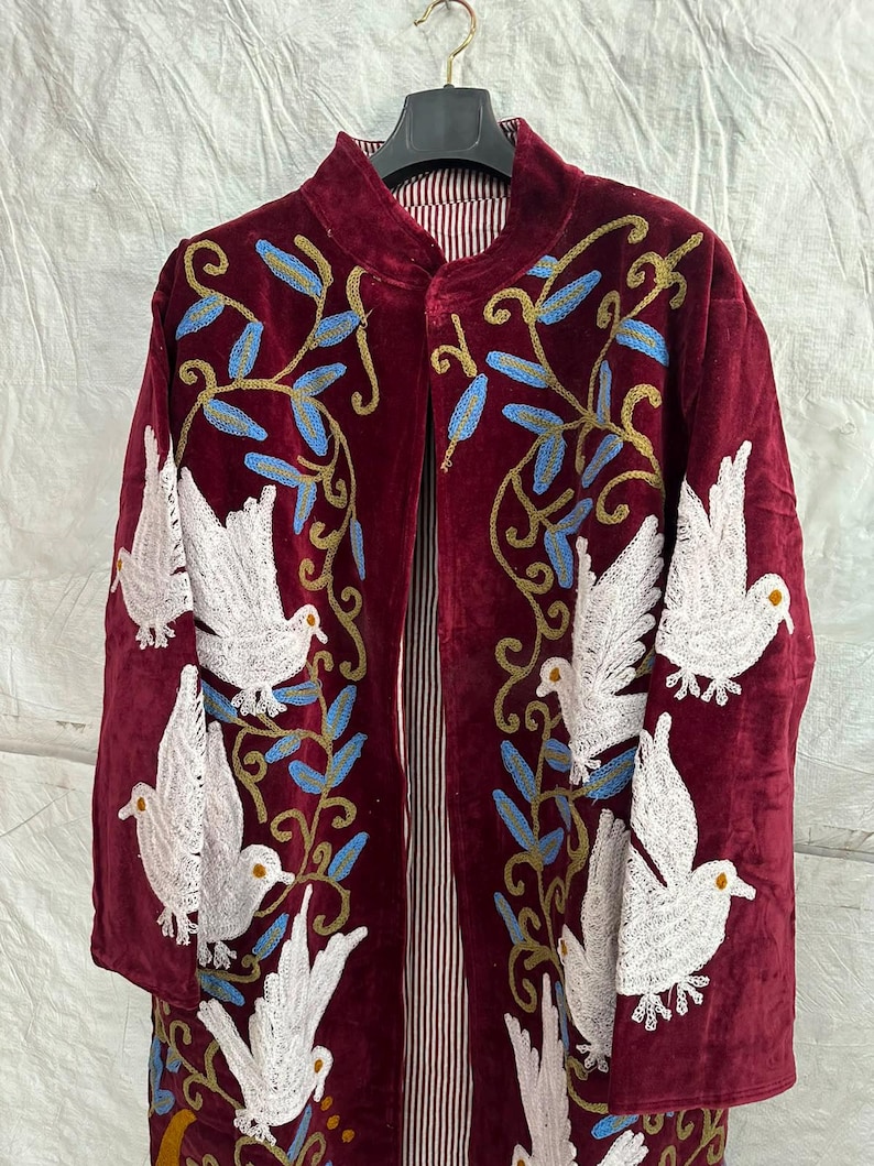 May include: A deep burgundy velvet coat featuring intricate embroidery of white doves and blue leafy vines. The garment has a high collar, long sleeves, and a vertical striped lining visible at the front opening, all displayed on a black hanger.