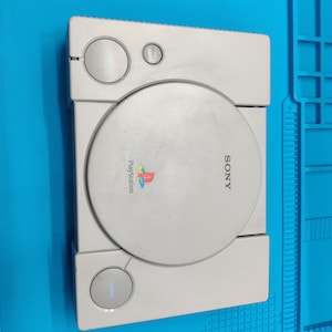 Może przedstawiać: Jasnoszara konsola do gier Sony PlayStation z okrągłą pokrywą napędu płyt, logo PlayStation i napisem Sony. Na górnej powierzchni znajduje się przycisk zasilania, przycisk resetowania oraz przycisk otwierania napędu w prostokątnej obudowie.