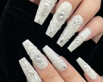May include: White, coffin-shaped artificial nails with intricate embellishments. Each nail features a combination of rhinestones, pearls, and delicate silver chains. The designs include ornate patterns and sparkling accents, creating a glamorous and elegant look.