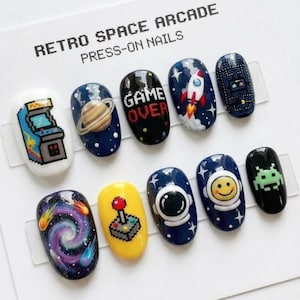 May include: A set of press-on nails with a retro space arcade theme. Designs include a pixelated arcade machine, planets, rockets, a joystick, and the words "GAME OVER". The nails are painted in various colors, including blue, yellow, and black.