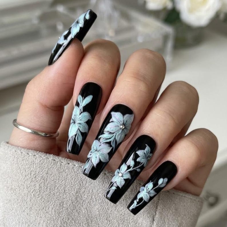 May include: Long coffin-shaped nails with a glossy black base coat. Each nail features hand-painted light blue floral designs with delicate white highlights and small silver metallic studs placed at the center of the flower petals.