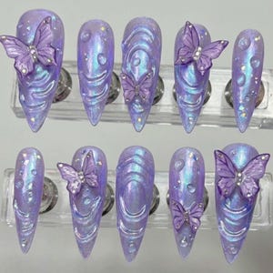 May include: A set of ten long, pointed artificial nails in a lavender iridescent color. Each nail features a 3D butterfly and water droplet design. The butterflies are a darker shade of purple and have rhinestone accents.
