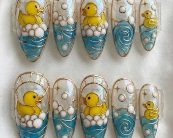 May include: A set of ten press-on nails featuring a yellow rubber duck design. Each nail showcases a yellow duck in a blue water scene with white bubbles and a gold outline. The nails are oval-shaped and have a glossy finish.