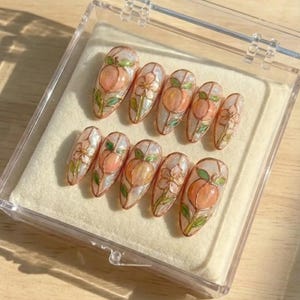 K&ouml;nnte beinhalten: Ein Set aus zehn k&uuml;nstlichen N&auml;geln mit Pfirsich- und Blumendesign. Jeder Nagel zeigt einen Pfirsich in Orange- und Rosat&ouml;nen, umgeben von gr&uuml;nen Bl&auml;ttern und wei&szlig;en Bl&uuml;ten. Die N&auml;gel werden in einer transparenten Kunststoffbox pr&auml;sentiert.