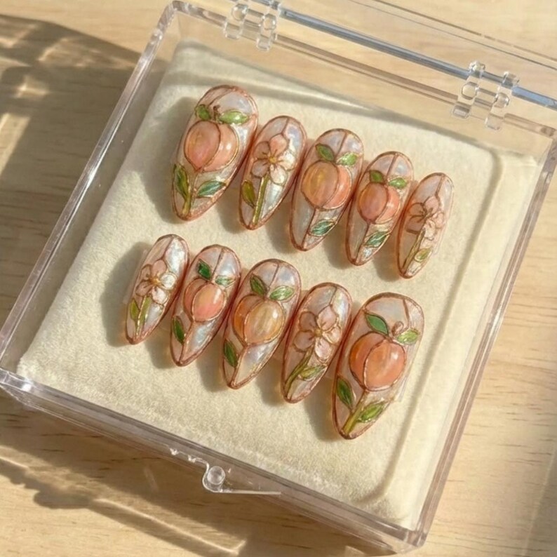 K&ouml;nnte beinhalten: Ein Set aus zehn k&uuml;nstlichen N&auml;geln mit Pfirsich- und Blumendesign. Jeder Nagel zeigt einen Pfirsich in Orange- und Rosat&ouml;nen, umgeben von gr&uuml;nen Bl&auml;ttern und wei&szlig;en Bl&uuml;ten. Die N&auml;gel werden in einer transparenten Kunststoffbox pr&auml;sentiert.