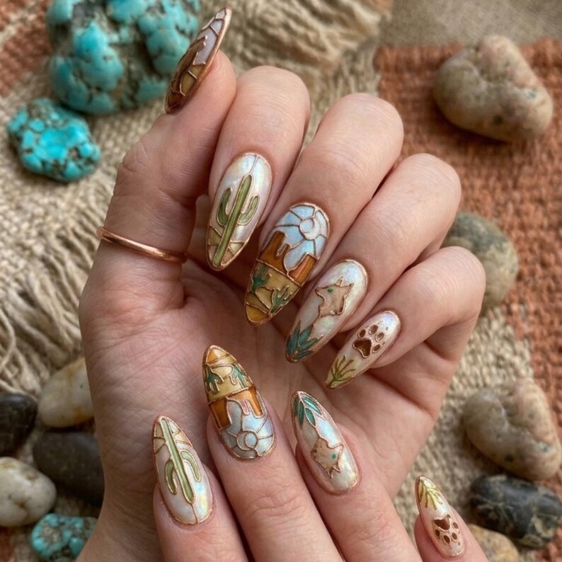 Puede incluir: Una mano con u&ntilde;as largas en forma de almendra con dise&ntilde;os de u&ntilde;as con tem&aacute;tica des&eacute;rtica. Los dise&ntilde;os incluyen cactus, un paisaje des&eacute;rtico, huellas de patas y una estrella. Las u&ntilde;as tienen una base nacarada con detalles dorados y marrones.
