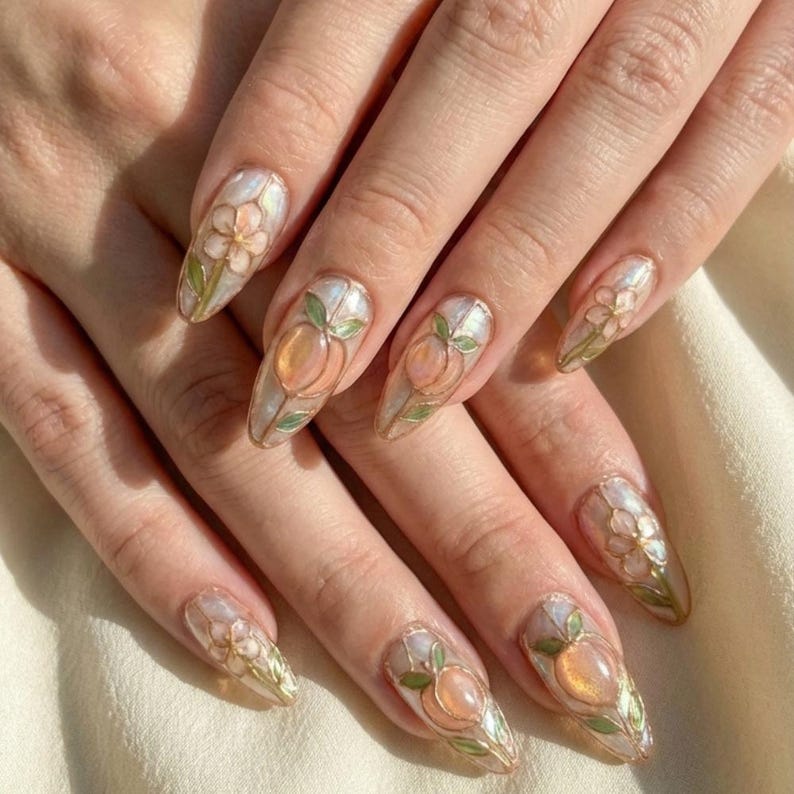 K&ouml;nnte beinhalten: Nahaufnahme von langen, mandelf&ouml;rmigen Fingern&auml;geln mit aufwendiger Nagelkunst. Das Design zeigt Pfirsich- und Blumenmotive in Pfirsich-, Gr&uuml;n- und Goldt&ouml;nen auf einer perlmuttartigen Basis. Die N&auml;gel sind gut gepflegt und zeigen einen zarten, k&uuml;nstlerischen Stil.