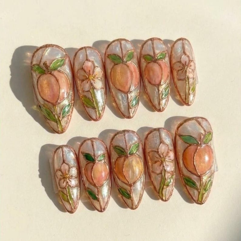 K&ouml;nnte beinhalten: Ein Satz von zehn mandelf&ouml;rmigen Kunstn&auml;geln. Jeder Nagel zeigt ein Pfirsich- oder Blumendesign mit gr&uuml;nen Bl&auml;ttern, umrandet von einer kupferfarbenen Bord&uuml;re. Die Pfirsichdesigns sind in Rosa- und Oranget&ouml;nen gehalten.