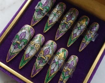 Può includere: Un set di dieci unghie finte lunghe e appuntite disposte in una scatola di velluto viola. Ogni unghia presenta un design dettagliato con un motivo a giglio in oro, verde e viola, bordato d'oro. Le unghie hanno un effetto vetrata.