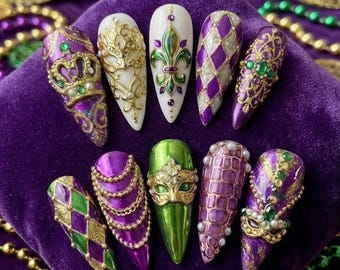 May include: A collection of ten long, almond-shaped artificial nails. The nails feature intricate designs in purple, gold, green, and white, including fleur-de-lis, harlequin patterns, and decorative masks. Some nails are adorned with pearls and jewels.