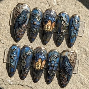 May include: A set of ten long, almond-shaped press-on nails. Each nail features a detailed design with a dark blue and bronze color scheme. Designs include ravens, flowers, and stained-glass lantern motifs. The nails are displayed on a neutral, textured surface.