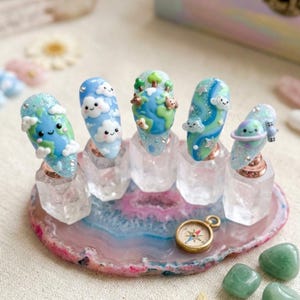 May include: A set of five Earth-themed nail art designs. Each nail features a different design, including clouds, the Earth, and planets. The nails are displayed on clear stands atop a pink and blue agate slice, with a compass and green stones.