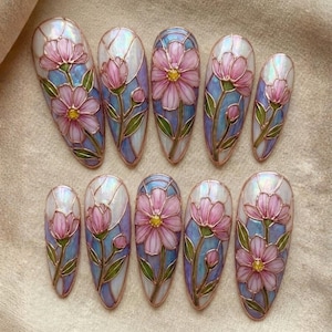 May include: A set of ten artificial nails with a floral design. Each nail features a pink flower with a yellow center, green leaves, and a blue and white background, all outlined in a copper color.