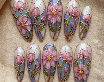 May include: A set of ten artificial nails with a floral design. Each nail features a pink flower with a yellow center, green leaves, and a blue and white background, all outlined in a copper color.