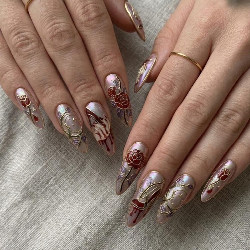 May include: Almond-shaped nails with detailed nail art. The design includes red roses, gold detailing, and a blood-like drip effect. The nails have a pearlescent base, creating a dramatic and artistic appearance.