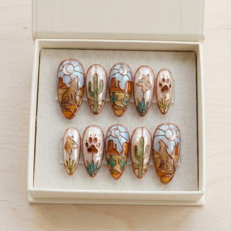 Puede incluir: Un juego de diez u&ntilde;as postizas en forma de almendra con dise&ntilde;os tem&aacute;ticos del desierto. Cada u&ntilde;a presenta un dise&ntilde;o diferente, que incluye un lobo aullando, cactus, huellas de patas y paisajes des&eacute;rticos en tonos marr&oacute;n, naranja, verde y azul. Las u&ntilde;as se presentan en una caja blanca.