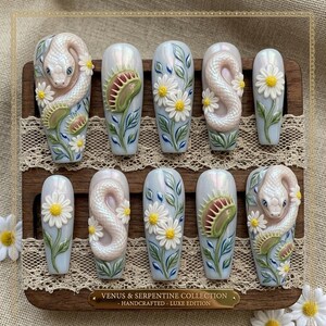 May include: A set of ten press-on nails with a light blue iridescent finish. Each nail features 3D designs of snakes, daisies, and Venus flytraps. The nails are displayed on a wooden stand with the text "VENUS & SERPENTINE COLLECTION - HANDCRAFTED - LUXE EDITION."