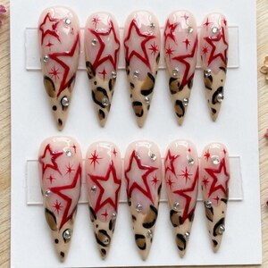 May include: Set of ten long, almond-shaped false nails. Each nail features a nude base with red star designs, small rhinestones, and leopard print accents. The nails are arranged on a white surface.