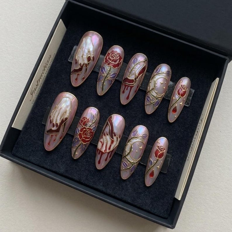 May include: A set of ten artificial nails with a pearlescent finish, each decorated with distinct designs. The designs include a red rose, a hand wearing a high heel, and a vine with leaves. The nails are presented in a black box.