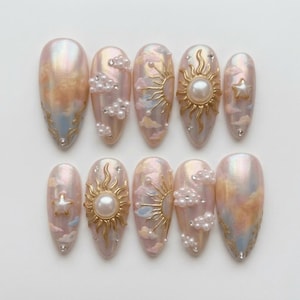May include: A set of ten almond-shaped artificial nails. The nails feature a pearlescent base with cloud and sun designs. Some nails have gold sun accents, pearl embellishments, and star details. The color palette includes peach, pink, and blue.