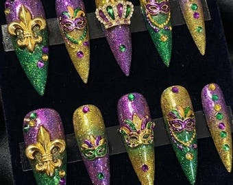 Può includere: Un set di dieci unghie finte lunghe e a punta con tema Mardi Gras. Le unghie presentano glitter verde, oro e viola, con abbellimenti come fleur-de-lis, maschere e una corona. Alcune unghie hanno accenti di gioielli.