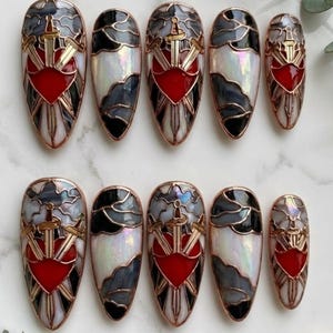 Puede incluir: Juego de diez uñas postizas con diseño de vidriera. Cada uña presenta un corazón rojo, espadas doradas y detalles grises y blancos, todo delineado en cobre. Las uñas tienen forma ovalada y un acabado brillante.