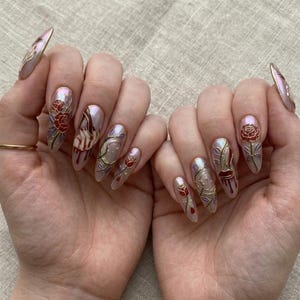May include: Almond-shaped artificial nails with a pearlescent sheen. Each nail showcases detailed designs, including red roses, gold vines, and abstract figures. The nails are finished with a glossy top coat, set against a neutral backdrop.