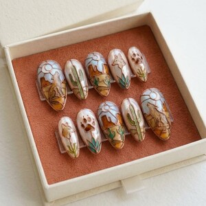 Puede incluir: Un conjunto de diez u&ntilde;as postizas ovaladas con dise&ntilde;os tem&aacute;ticos del desierto. Cada u&ntilde;a presenta un dise&ntilde;o diferente, que incluye un lobo aullando, cactus, paisajes des&eacute;rticos y huellas de patas, todo en tonos marrones, verdes y azules.