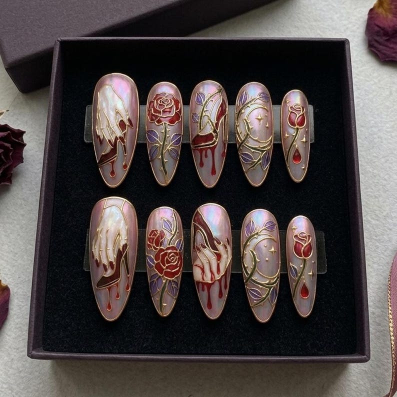 May include: A set of ten press-on nails in a dark box. Each nail features a design with a hand, a rose, and vines in red, gold, and purple on a pearlescent pink background. The nails are oval-shaped.