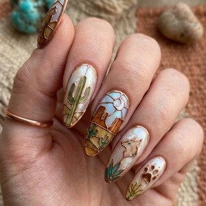 Puede incluir: Mano con u&ntilde;as largas y ovaladas con dise&ntilde;os tem&aacute;ticos del desierto. Las u&ntilde;as muestran un cactus, un paisaje des&eacute;rtico y un lobo. Los dise&ntilde;os est&aacute;n delineados en cobre y rellenos con esmalte blanco, verde, marr&oacute;n y azul.