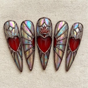 Puede incluir: Cinco piezas de arte de uñas de vidrieras. Cada uña presenta una forma de corazón rojo, enmarcada por paneles de vidrio iridiscente. Las uñas tienen forma puntiaguda y están dispuestas sobre un fondo de tela neutra.