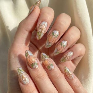 K&ouml;nnte beinhalten: Mandelf&ouml;rmige Nagelkunst mit Pfirsich- und Blumendesigns. Die N&auml;gel haben eine perlmuttartige Basis mit pfirsichfarbenen Fr&uuml;chten und wei&szlig;en Bl&uuml;ten. Gr&uuml;ne Bl&auml;tter und goldene Umrisse vervollst&auml;ndigen das Design. Die N&auml;gel sind lang und gepflegt.