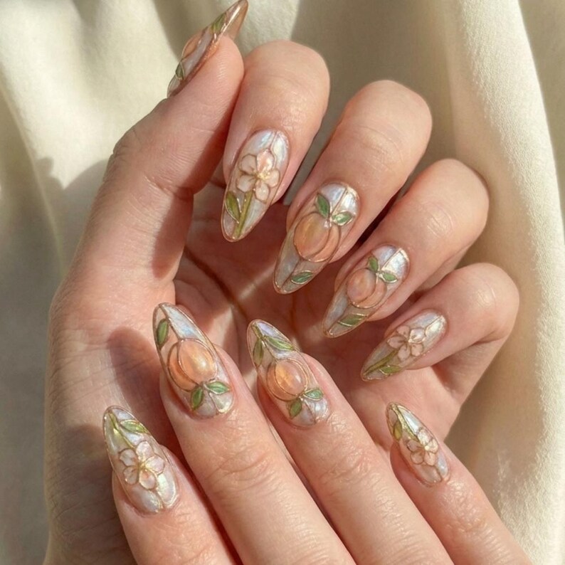 K&ouml;nnte beinhalten: Mandelf&ouml;rmige Nagelkunst mit Pfirsich- und Blumendesigns. Die N&auml;gel haben eine perlmuttartige Basis mit pfirsichfarbenen Fr&uuml;chten und wei&szlig;en Bl&uuml;ten. Gr&uuml;ne Bl&auml;tter und goldene Umrisse vervollst&auml;ndigen das Design. Die N&auml;gel sind lang und gepflegt.