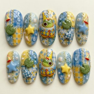 May include: A set of ten artificial nails with a whimsical, aquatic theme. Each nail features a unique design, including a frog in a crown, stars, water droplets, and lily pads. The color palette includes blue, yellow, and white, with a patchwork effect.