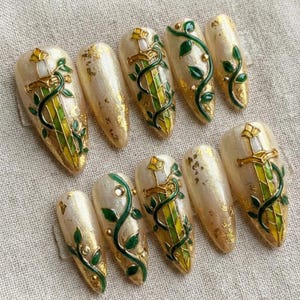 May include: Set of ten long, almond-shaped artificial nails. Each nail features a cream base with gold leaf accents. Designs include gold swords, green vines, and yellow and green stained glass patterns.